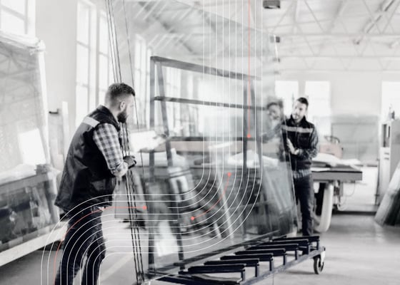 Workers handling large glass panels inside a manufacturing facility, representing glass production, supplier coordination, shipment visibility, and supply chain orchestration.